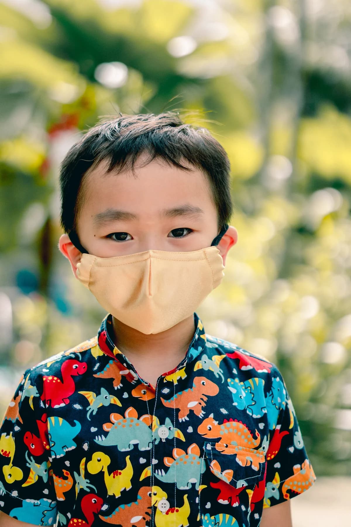 Young boy in a dinosaur shirt wearing a peach PEFORE. mask